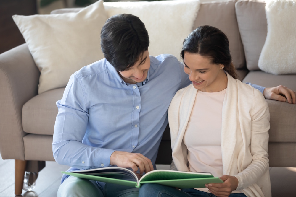 Couple Looking at Photobook Together Next to Sofa at Home Couple sitting next to a sofa looking at a personalised photobook together