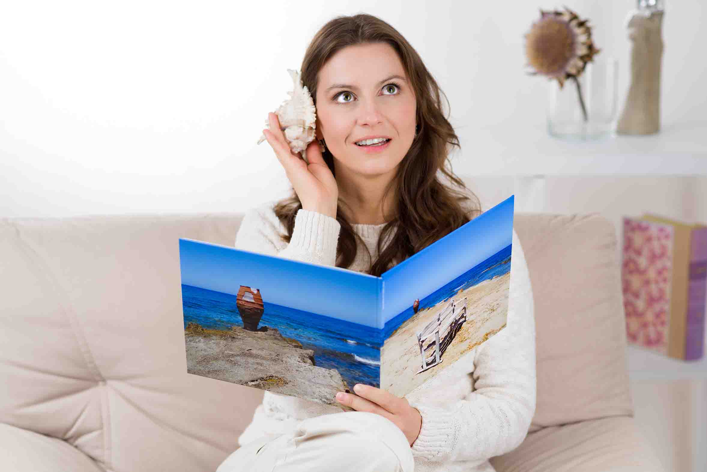 Woman looking through a square photobook while holding a seashell, enjoying printed travel memories in a personalised photo book.